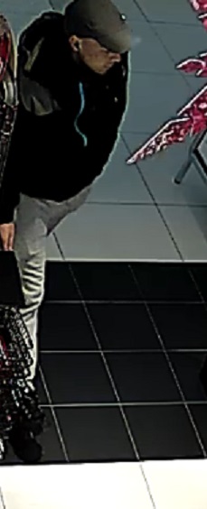 mężczyzna w wieku ok 25-30 lat, szczupłej budowy ciała, średniego wzrostu, ubrany w czarną kurtkę z kapturem, szarą czapkę z daszkiem, dresowe spodnie - koloru jasnego, na pasie ma przewieszoną czarną saszetkę marki Adidas