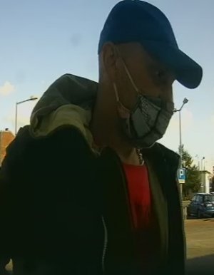 mężczyzna ma zakryte usta i nos maseczką ochronną. ma czapkę z daszkiem, blond włosy i koszulkę typu t-shirt koloru czerwonego a na niej bluzę z kapturem.
