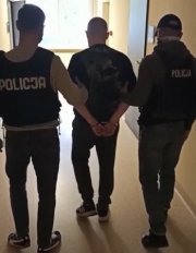 mężczyzna stojący na korytarzu komendy, tyłem w asyście dwóch policjantów kryminalnych. Mężczyzna ma założone kajdanki na ręce trzymane z tyłu.