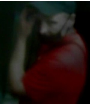 To mężczyzna tęgiej budowy ciała w koszulce koloru czerwonego z krótki rękawem, w czarnej czapce z daszkiem oraz krótkich spodenkach. Przez ramię ma przewieszoną torbę.