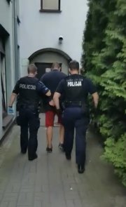 dwóch policjantów w umundurowaniu prowadzi zatrzymanego, który ma założone kajdanki na ręce trzymane z tyłu. Idą do drzwi wejściowych do komisariatu