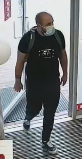 mężczyzna z widoczną łysiną na głowie i zakolami, na twarzy ma maseczkę zakrywającą usta i nos, ubrany w czarny tshirt, czarne dresowe spodnie i plecak