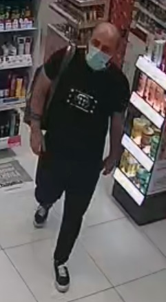 mężczyzna z widoczną łysiną na głowie i zakolami, na twarzy ma maseczkę zakrywającą usta i nos, ubrany w czarny tshirt, czarne dresowe spodnie i plecak