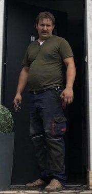 mężczyzna postawnej budowy ciała, ma średniej długości brązowe włosy, wąsa, ubrany w strój pracowniczy, spodnie granatowe oraz koszulkę z krótkim rękawem w kolorze oliwkowym, buty brązowe