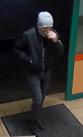 mężczyzna w wieku ok 30 -35 lat, szczupłej budowy ciała, ubrany w szarą zimową czapkę, zieloną, krótką, zimową kurtkę czarne spodnie dżinsy i sportowe buty, na twarzy ma komin zakrywający usta i nos