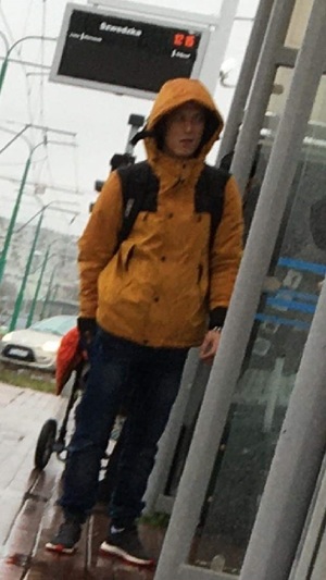 mężczyzna ubrany w pomarańczową kurtkę  z kapturem na głowie, ciemnym plecakiem na plecach, ciemnych spodniach i sportowych butach