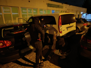 Policjanci stoją nad siedzącym i zakutym w kajdanki zatrzymanym. Jeden policjant kuca przy podejrzanym i z nim rozmawia. Osoby stoją przy zabezpieczonym Mercedesie.